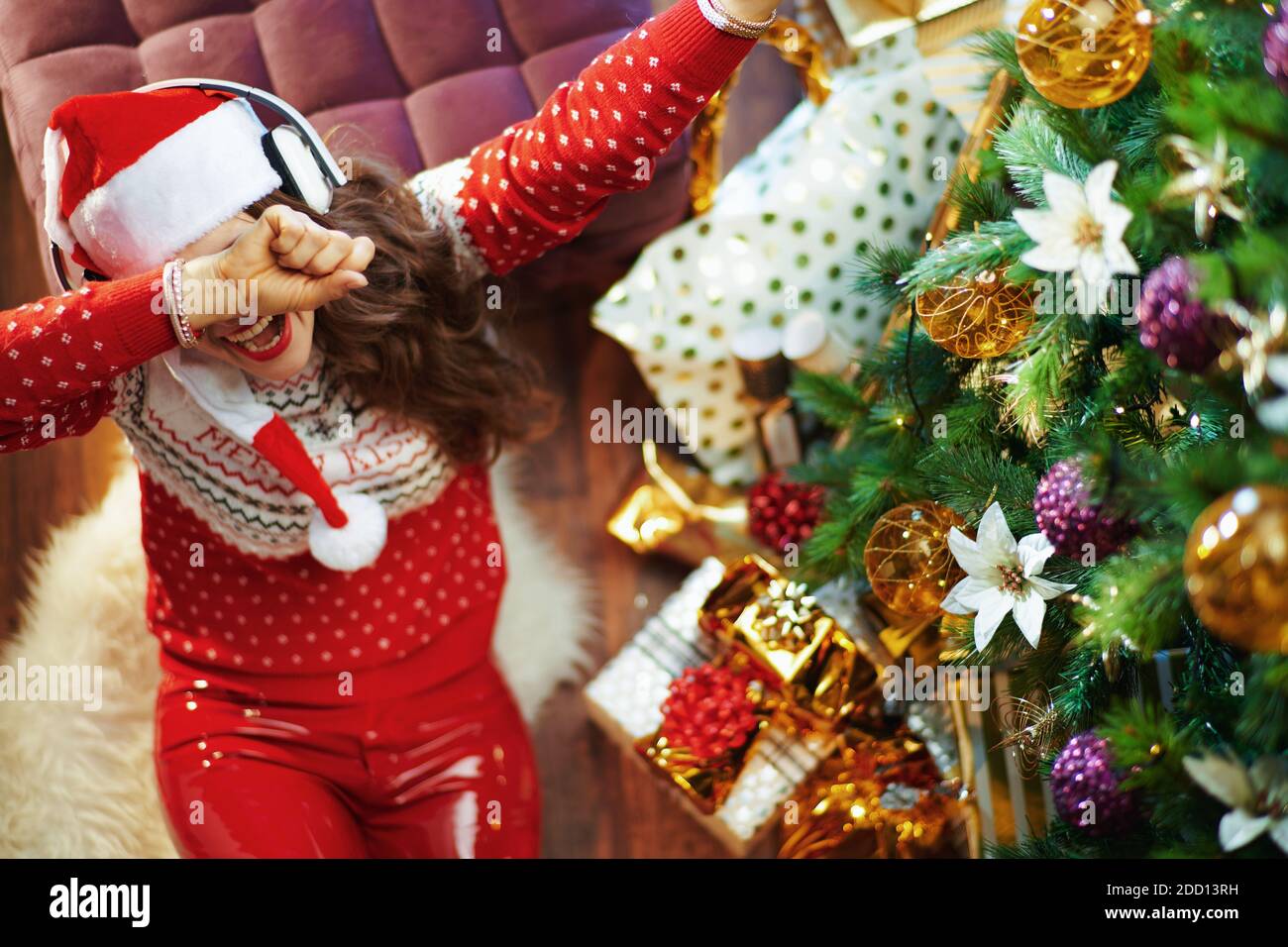 Merry Christmas. Upper view of happy young woman in red sweater and Santa hat listening to the music with headphones and doing dabbing gesture near Ch Stock Photo