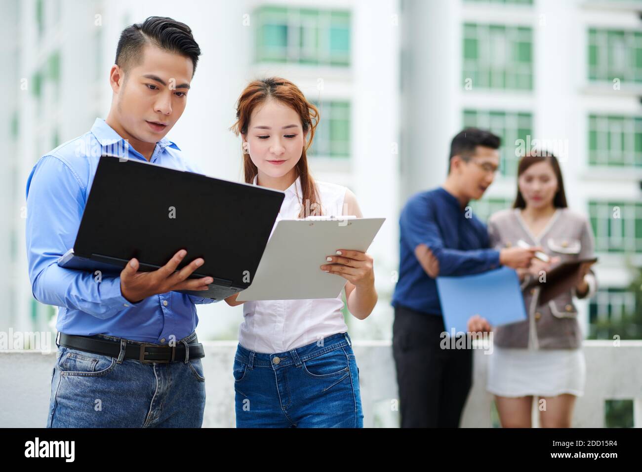 Young Vietnamese male and female software developers comparing programming code on tablet and laptop when standing outdoors Stock Photo