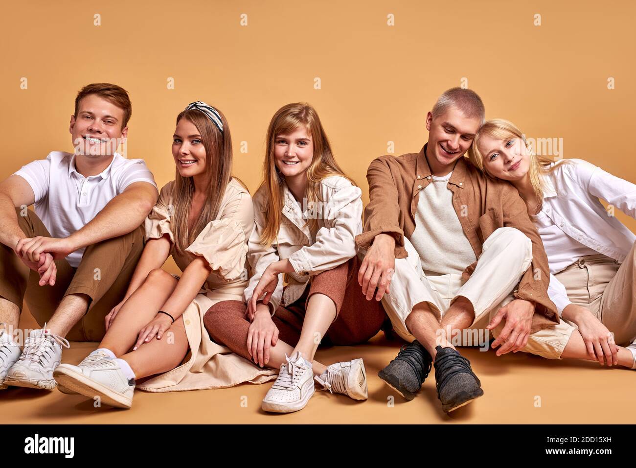 friendly group of students youth on the floor in studio on brown background, men and women in beige brown clothes posing together, as one team Stock Photo
