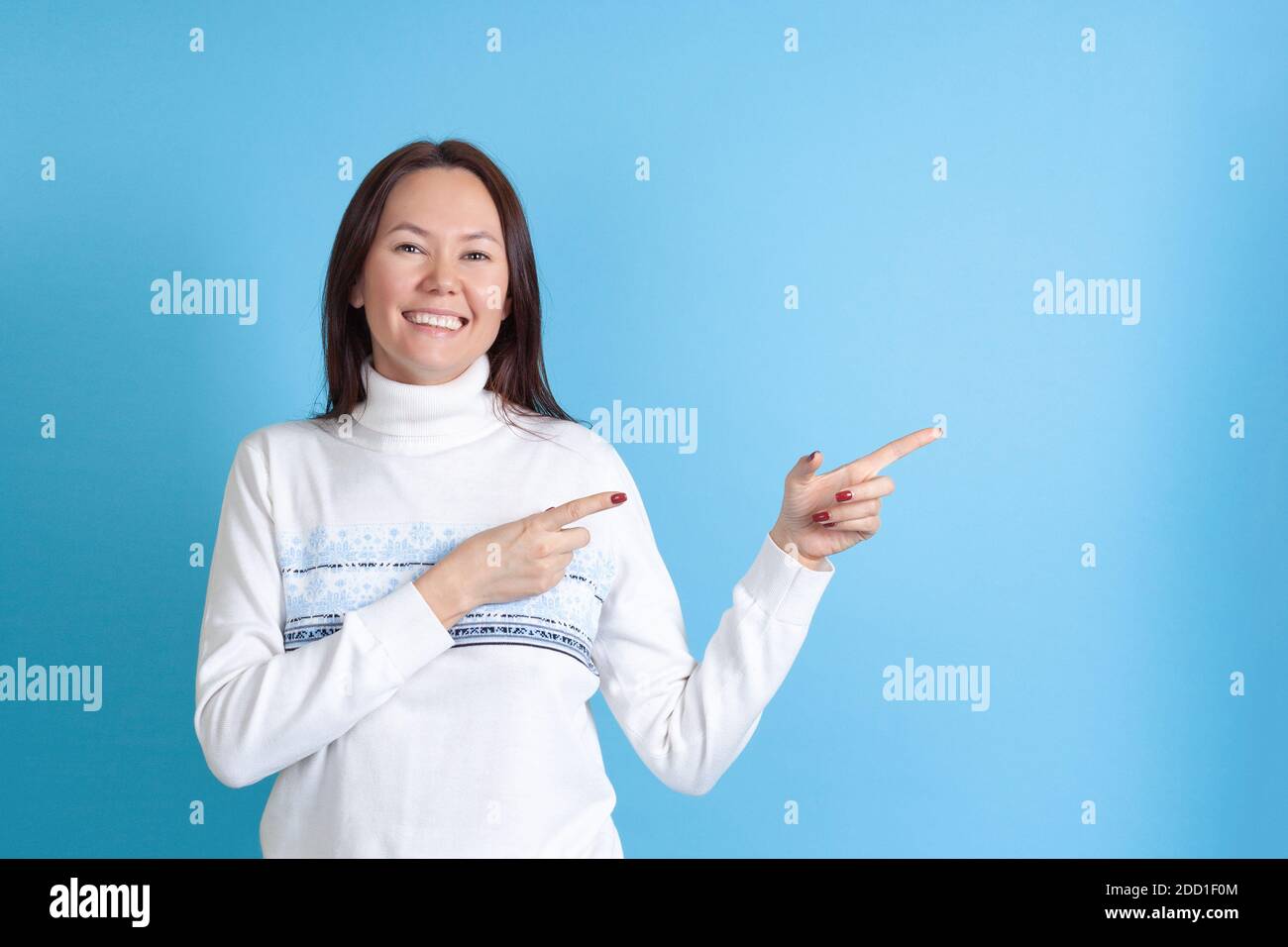 mock up a smiling Asian woman in a Christmas sweater points with her index fingers at an empty space for text isolated on a blue background Stock Photo