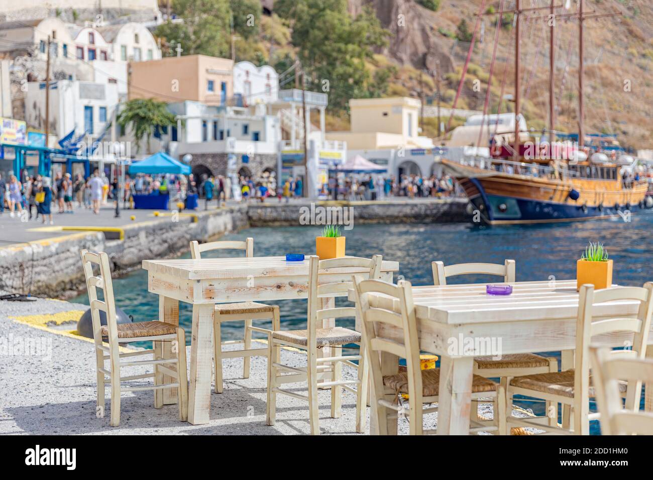 Chairs and tables of a traditional Greek taverna, restaurant at the dock of small port of blurred sail boat and tourist waking and shops. Santorini Stock Photo