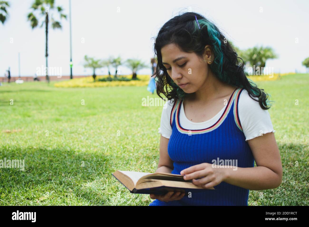 A young female sitting on the green grass in a park and reading a book ...