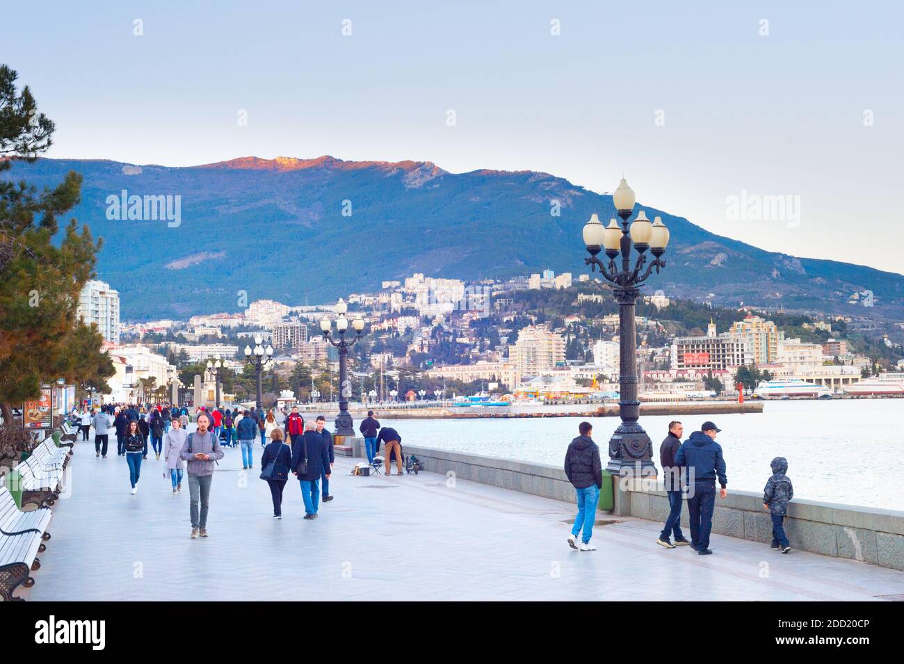 YALTA, CRIMEA - APRIL 3, 2018: People walking by resort city embankment, mountain in sunset light in background Stock Photo