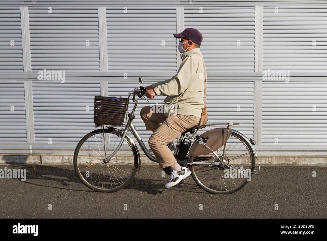 An older Japanese man, wearing a face mask, rides a bicycle in front of a metal wall in rural Kanagawa, Japan Stock Photo