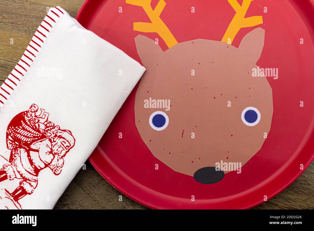 A red disposable plate with a funny rein deer figure inside is placed on top of a wooden table. A white cloth napkin with Santa Claus image on is plac Stock Photo