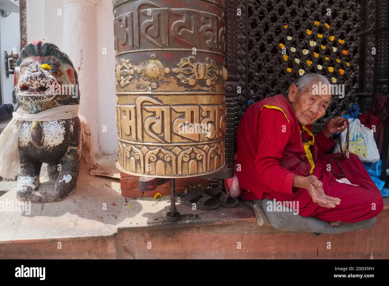 An elderly female Tibetan pilgrim begging for alms next to a large prayer wheel at the foot of the giant Bodhnath Stupa (Baudha) in Kathmandu, Nepal Stock Photo