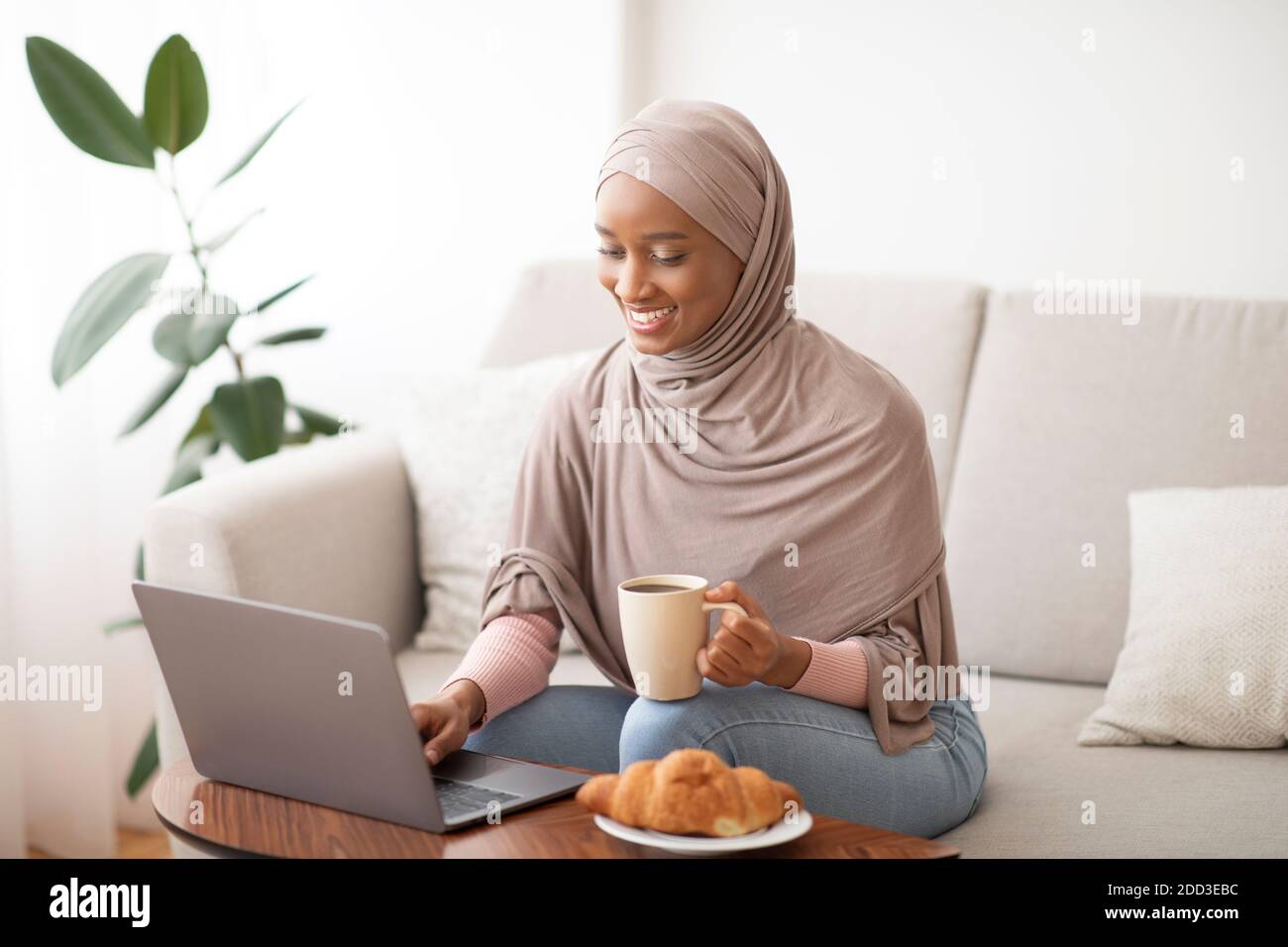 Online work, education, communication concept. Happy black lady in hijab using laptop and drinking coffee at home Stock Photo