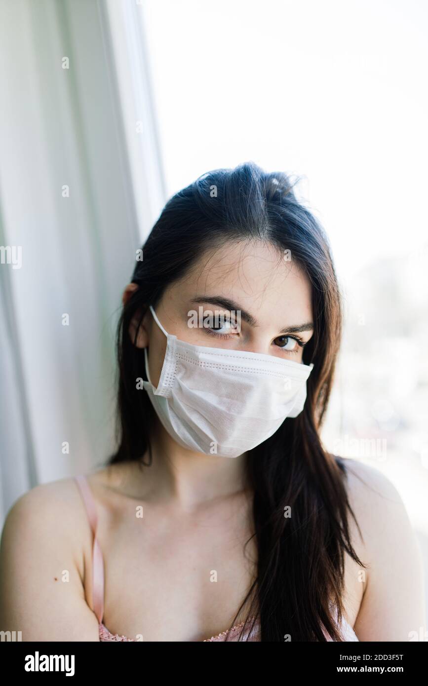 Lonely female isolated at home in protective medical mask on her face looking at window, bored because of pandemic coronavirus, covid. Stock Photo