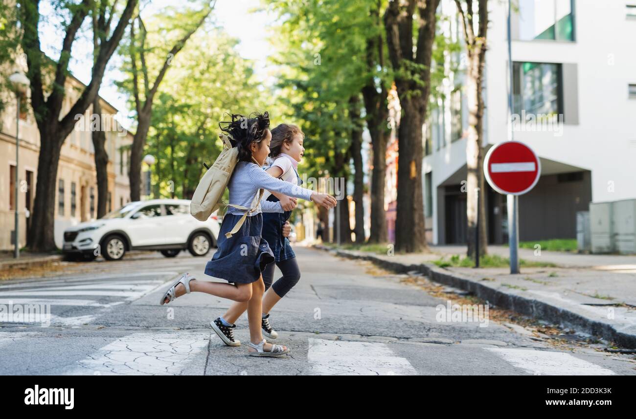 Side view of small school girls crossing street outdoors in town, running. Stock Photo