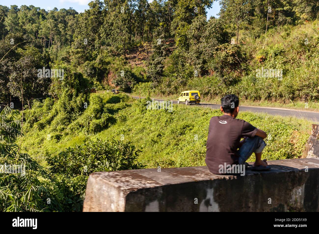 A man sits on the protective guarding next to a highway watching the traffic drive by him. A Tata Sumo SUV is visible in the picture. Stock Photo
