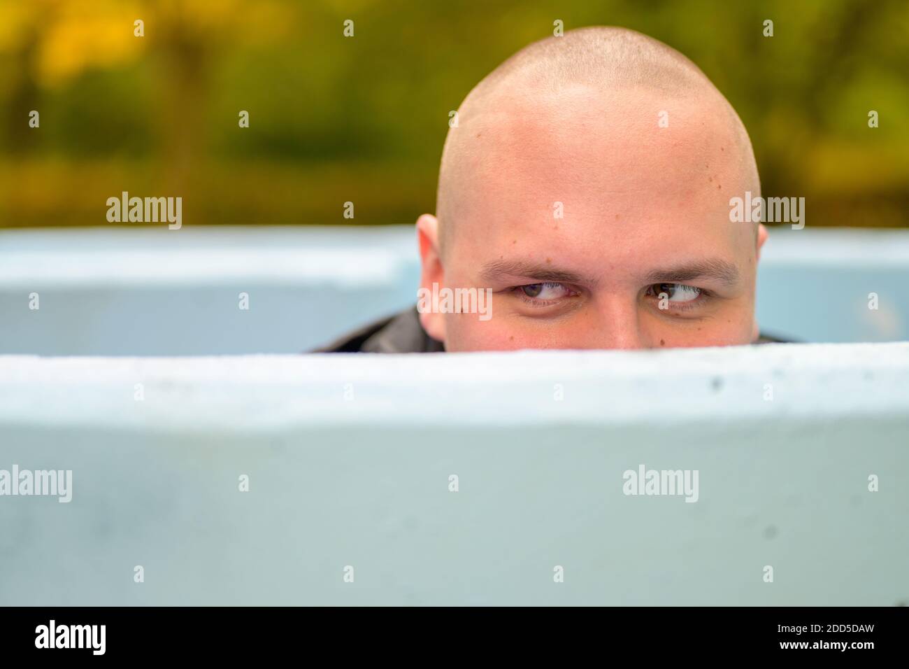 Young man with shaved head hunkering down in an outdoor tub or pool peering over the rim glancing to the side Stock Photo