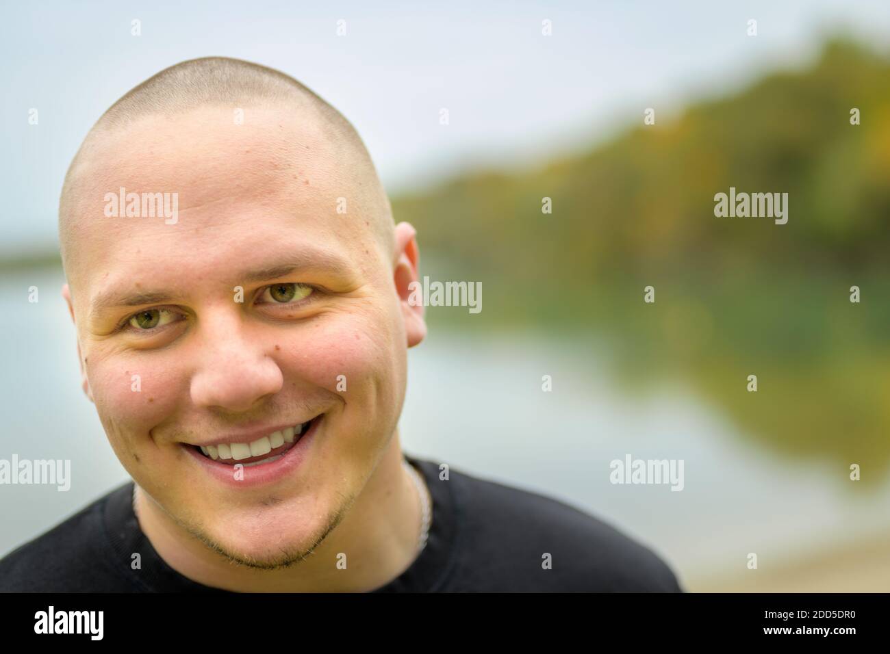 Unshaven young man with a lovely warm friendly smile in a close up head portrait looking to the side of the frame with tranquil lake background Stock Photo