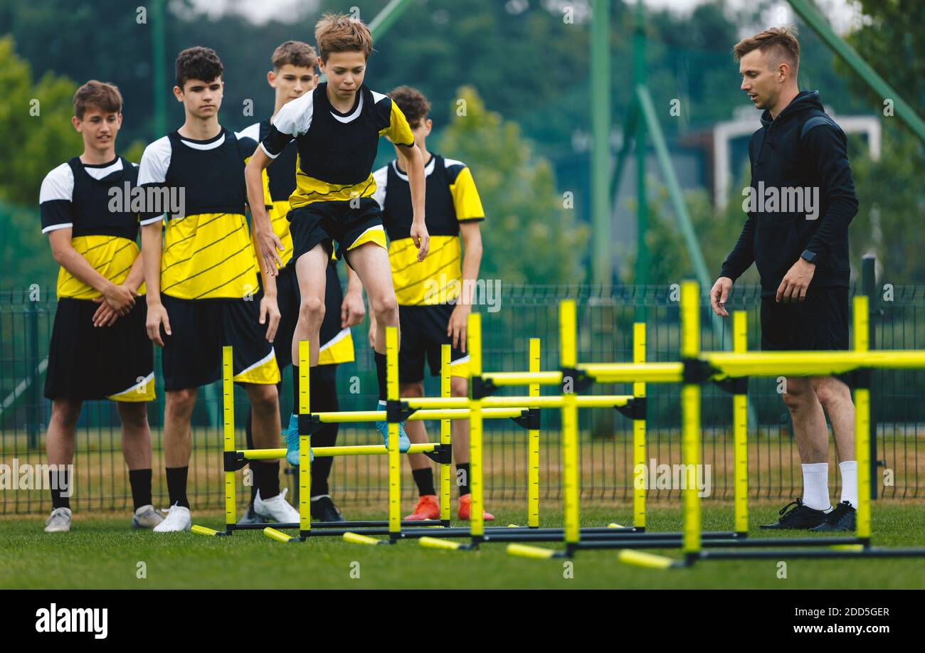 Boy Jumping Over Hurdles On Soccer Training. Football School Team On Training with Young Coach. Teens On Physical Education Class Stock Photo