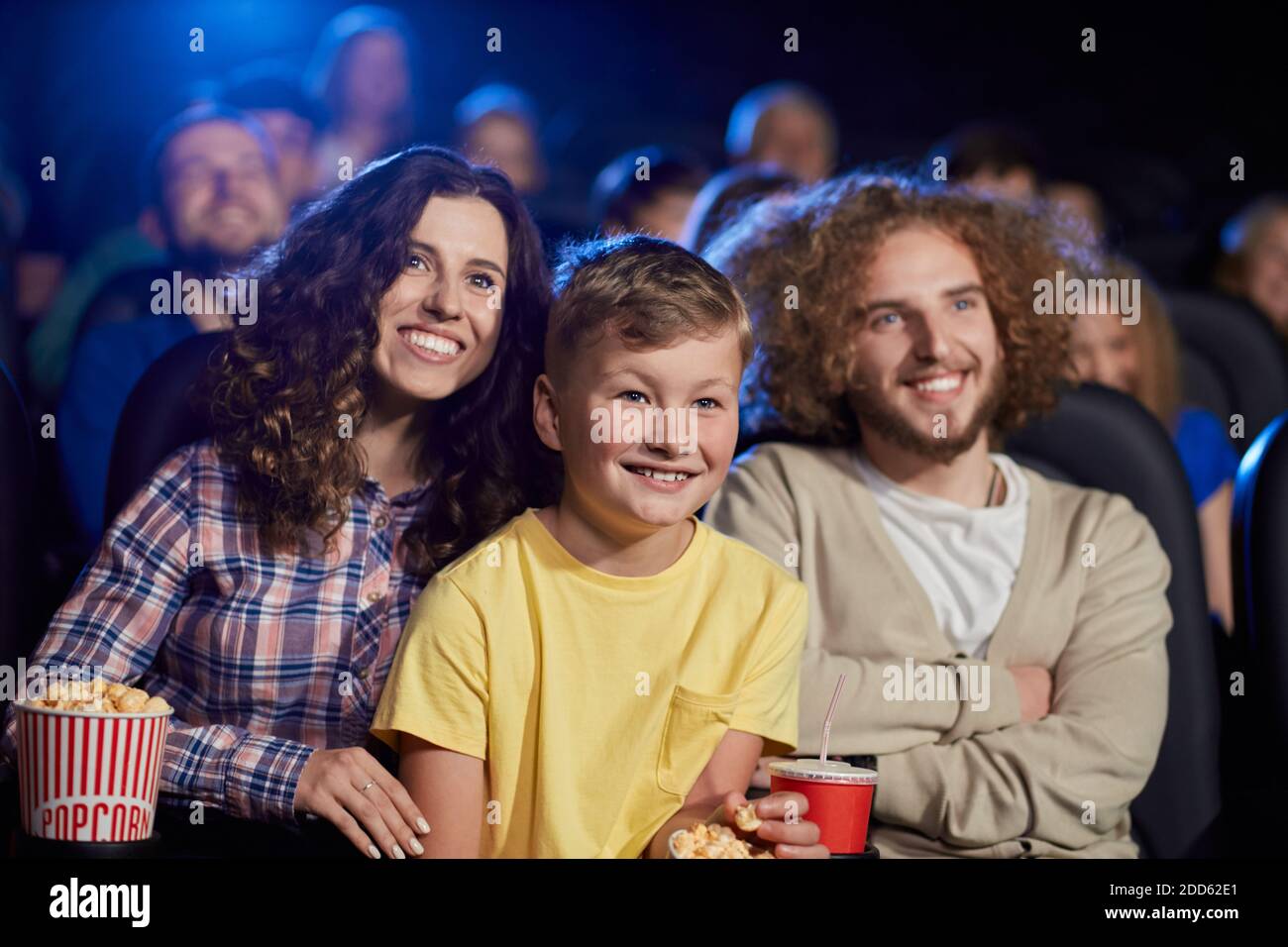 Selective focus of young parents with curly hair enjoying time with son holding sparkling drink, audience on background. Happy family sitting in cinema, kid on knees, watching funny cartoon. Stock Photo