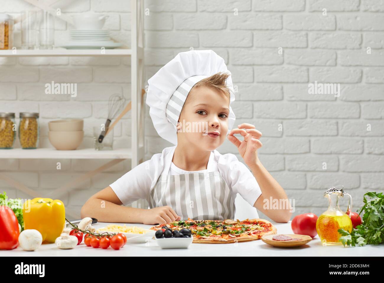 child making tasty delicious gesture by kissing fingers. little boy in chef hat and an apron cooking pizza in the kitchen. Stock Photo