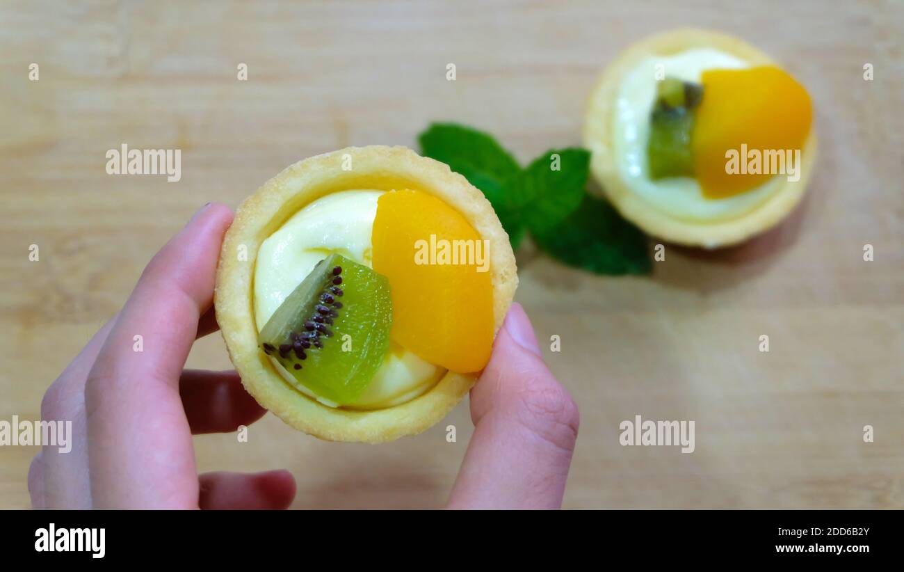 A hand holding a mini fruit tart, with another on the table in the blurred background. Stock Photo