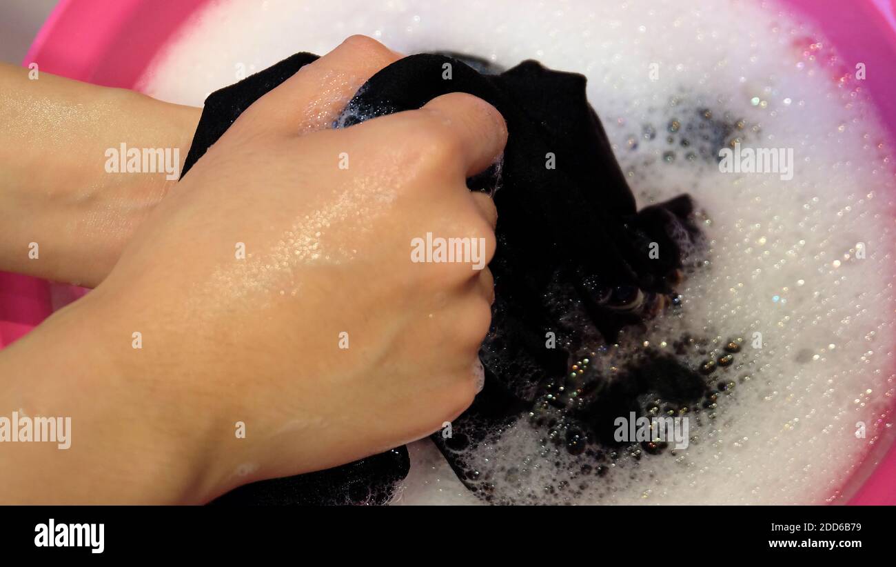Hands washing a black clothing in soapy water with bubbles, in a pink basin. Stock Photo