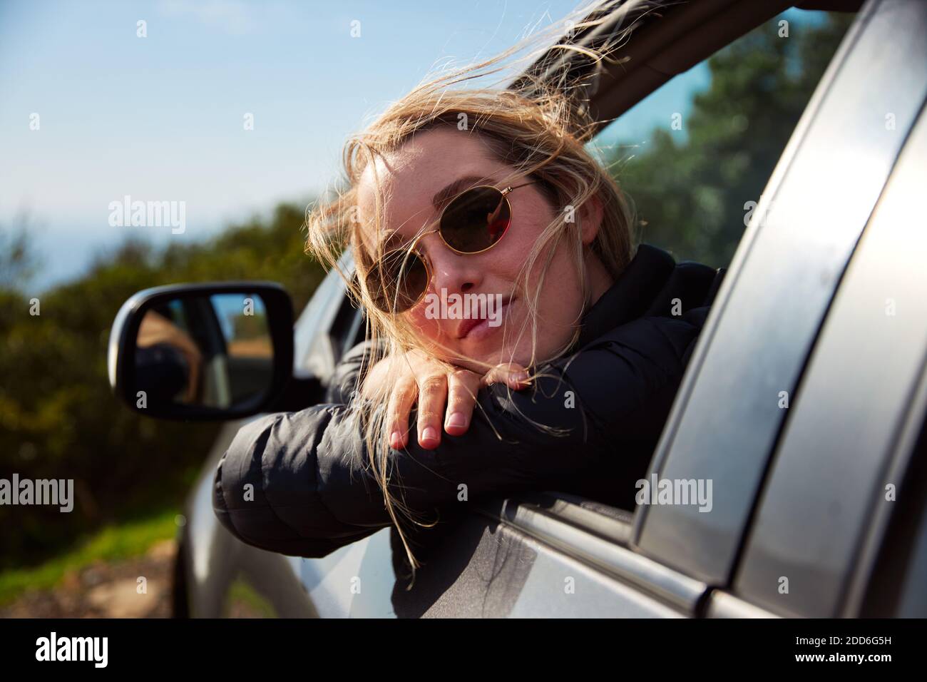 Young woman wearing sunglasses with eyes closed on road trip vacation leaning out of rental car window with beautiful mountain landscape behind Stock Photo