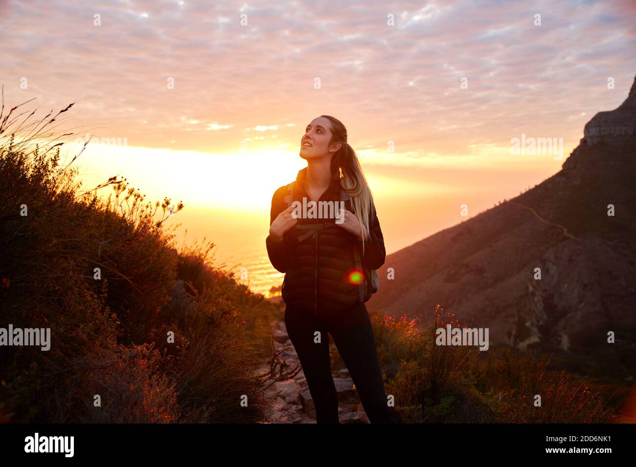 Front view of young woman on vacation wearing backpack setting off on hike climbing coastal path at sunset Stock Photo