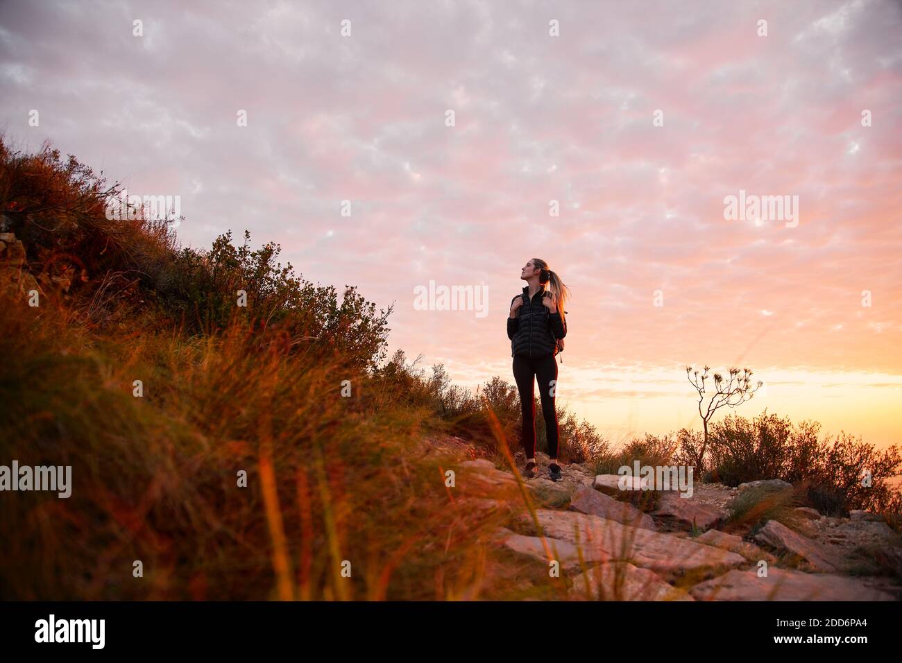 Front view of young woman on vacation wearing backpack setting off on hike climbing countryside path at sunset Stock Photo