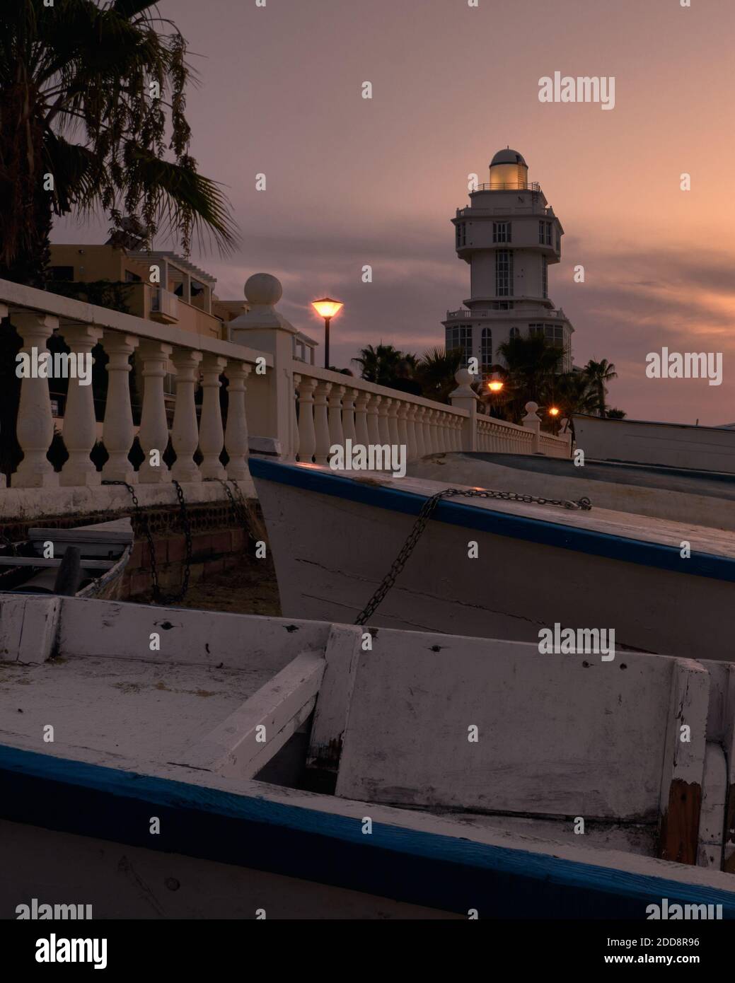 Faro de Isla Cristina al atardecer, en los alrrededores un paseo con farolas y unas barcas. Stock Photo