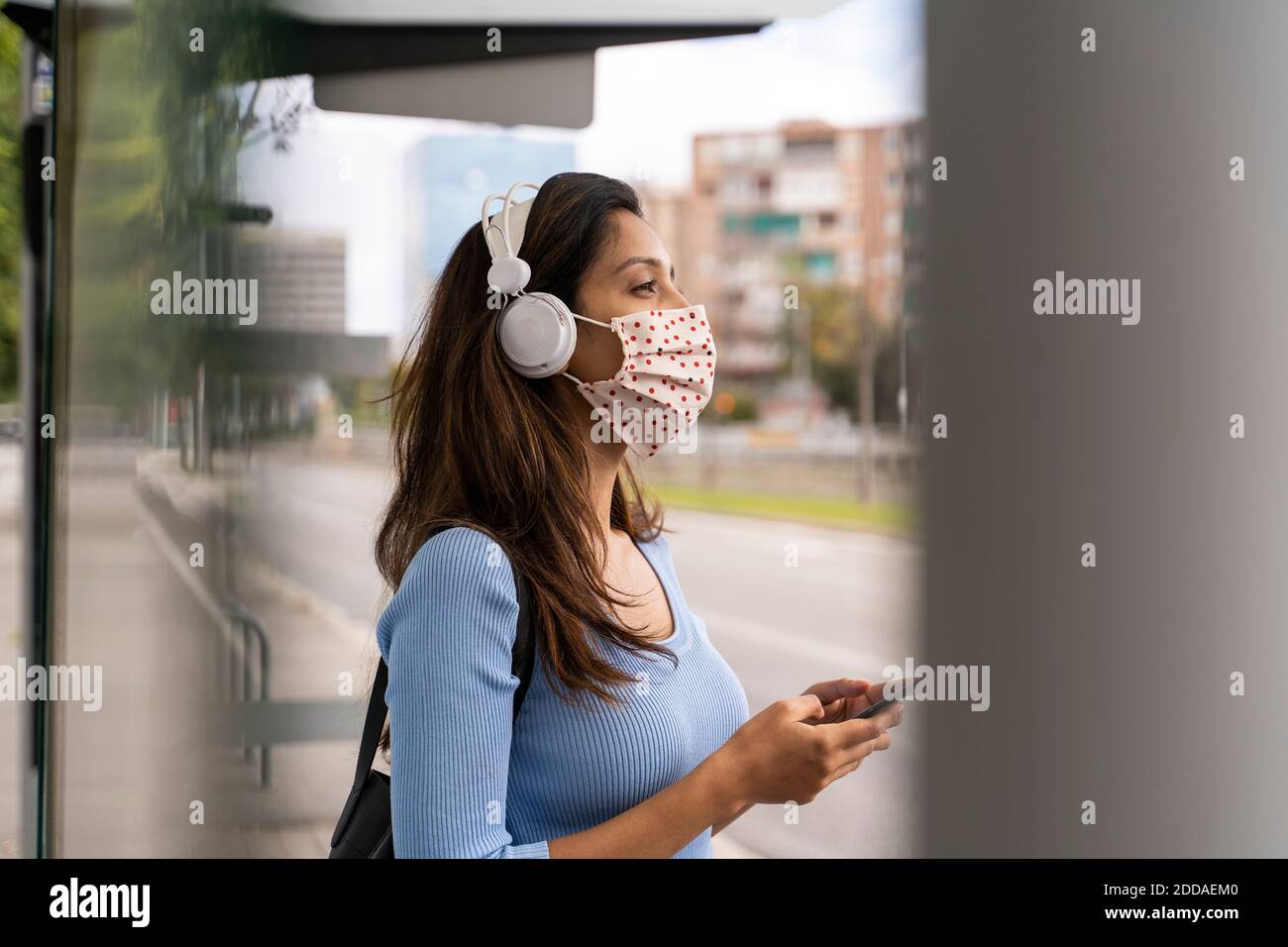 Contemplating woman in face mask wearing headphones with smart phone at bus stand during COVID-19 Stock Photo