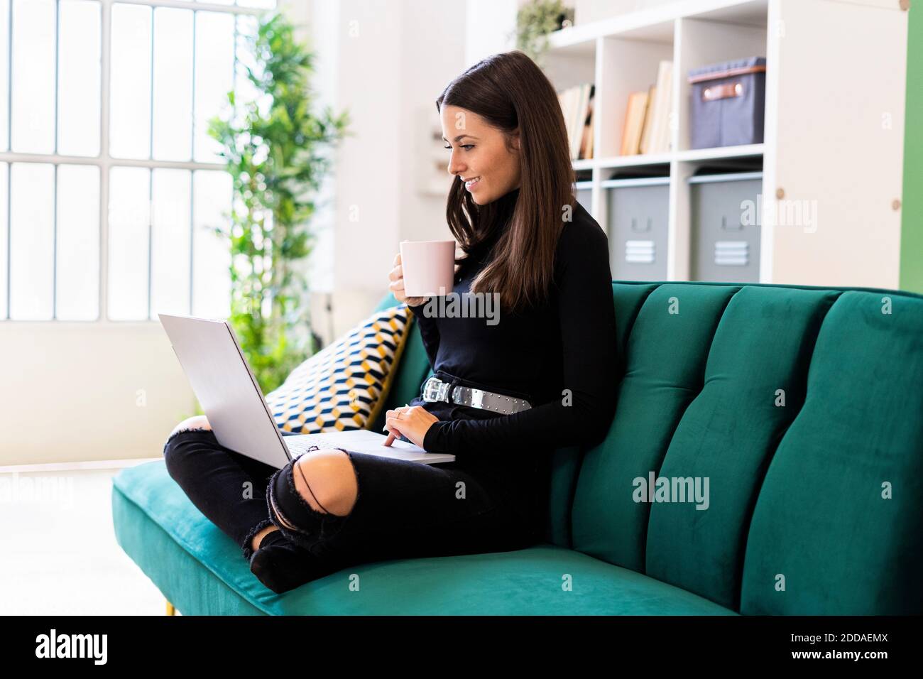 Smiling young woman holding coffee cup while using laptop for blogging at home Stock Photo