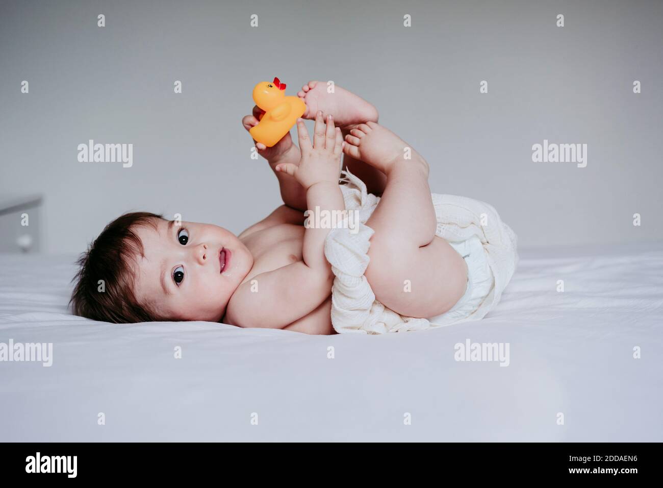 Cute baby boy playing with duck toy while lying down on bed at home Stock Photo