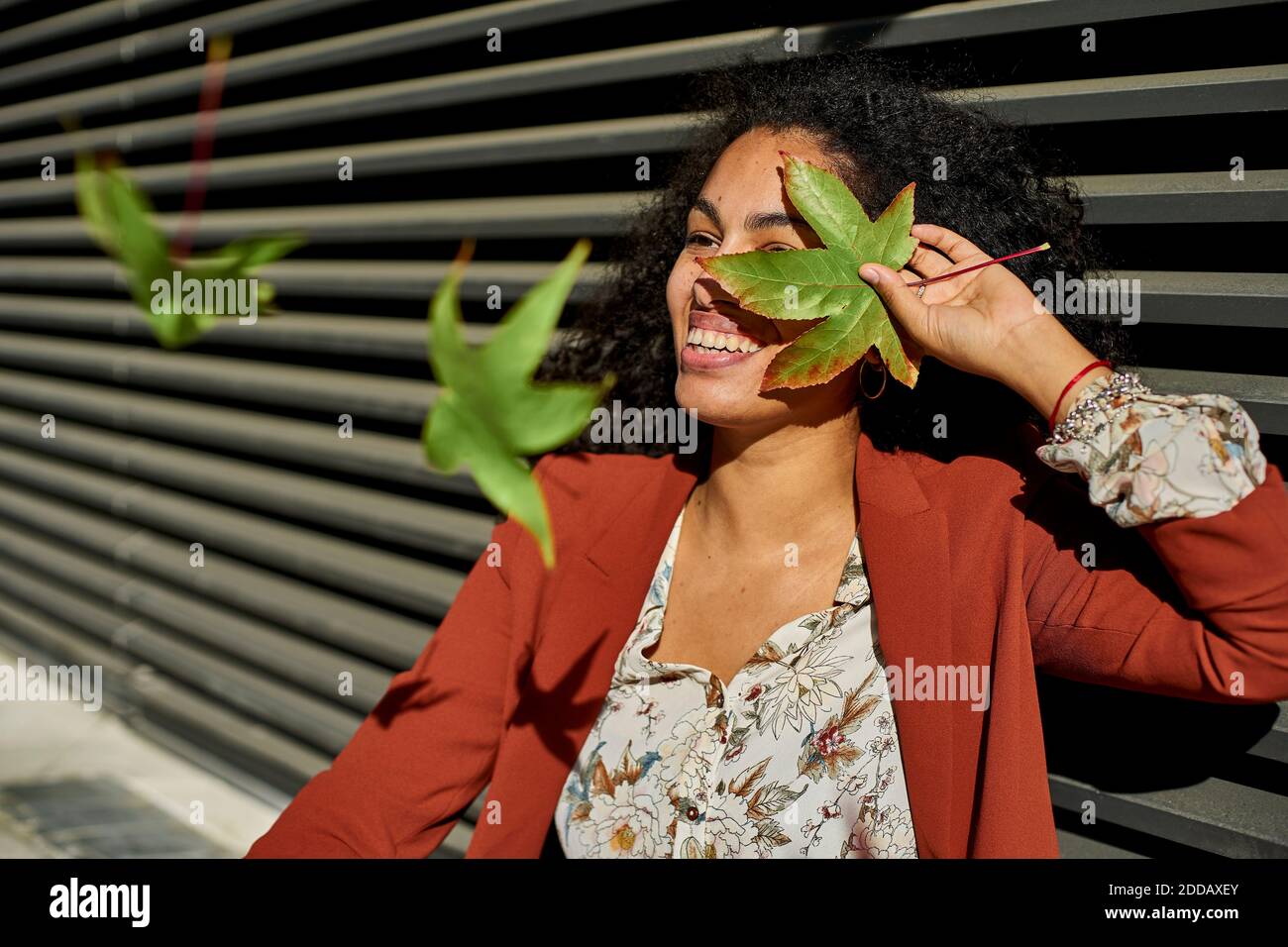 Autumn leaves falling on smiling young woman against shutter during sunny day Stock Photo