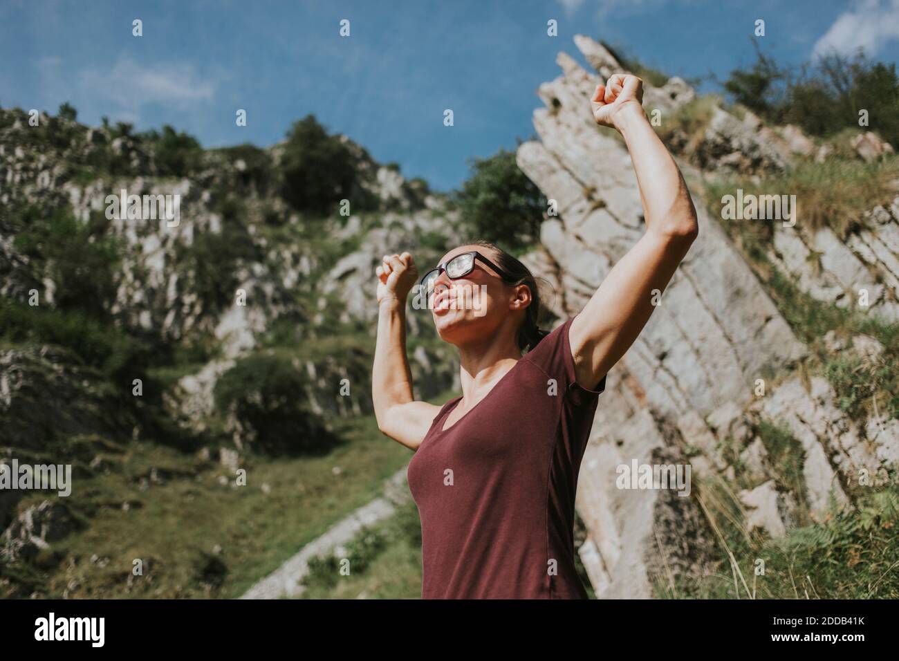 Woman celebrating victory with arms raised against mountain on sunny day Stock Photo