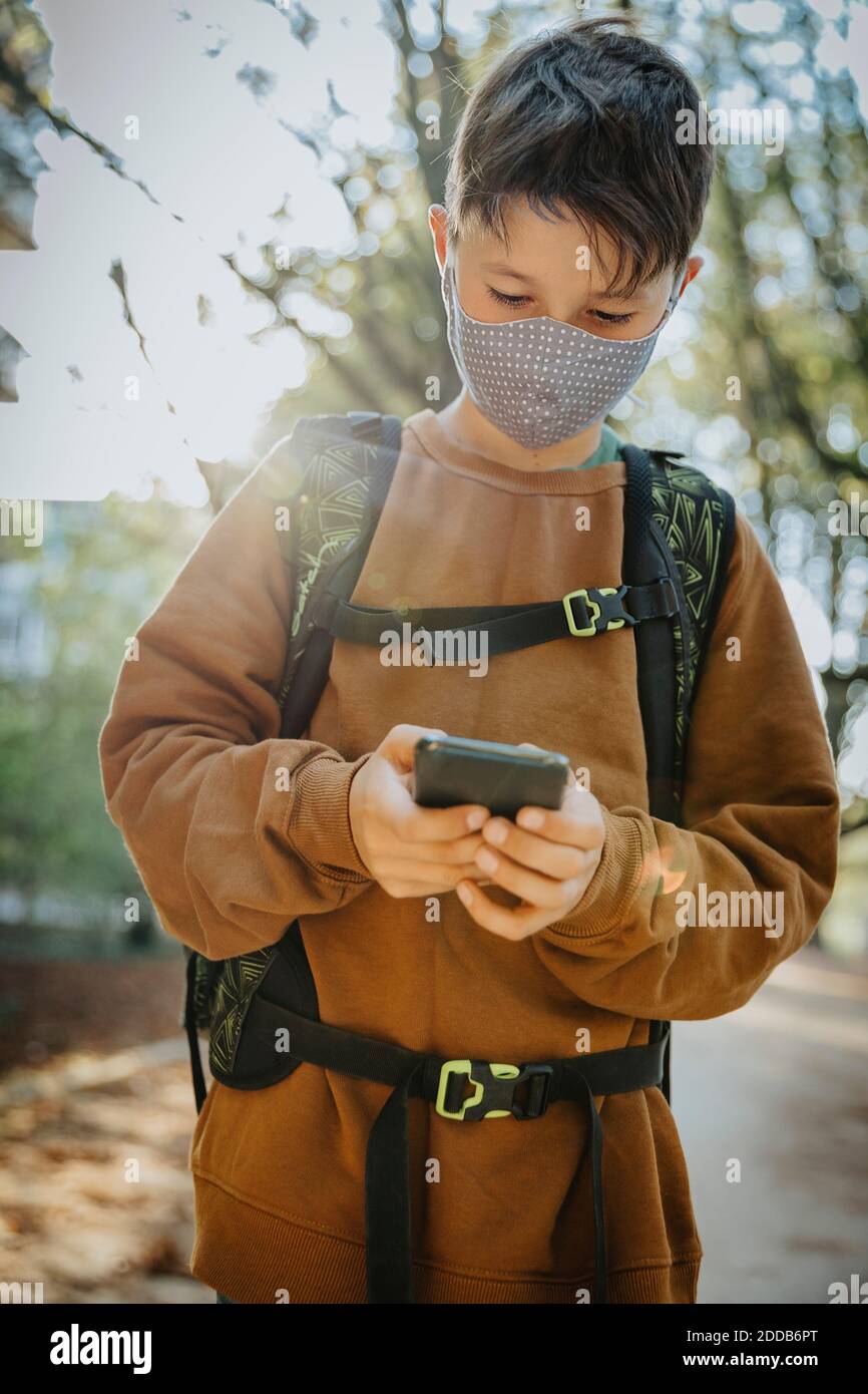Boy using smart phone wearing protective face mask while standing in public park Stock Photo