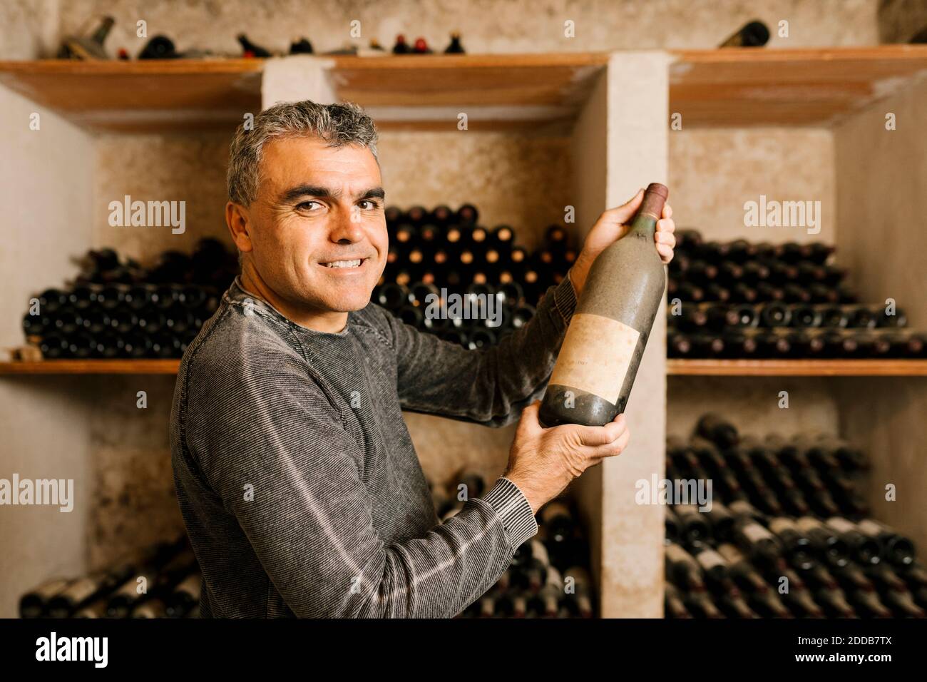 Smiling male owner holding old wine bottle while standing by rack at