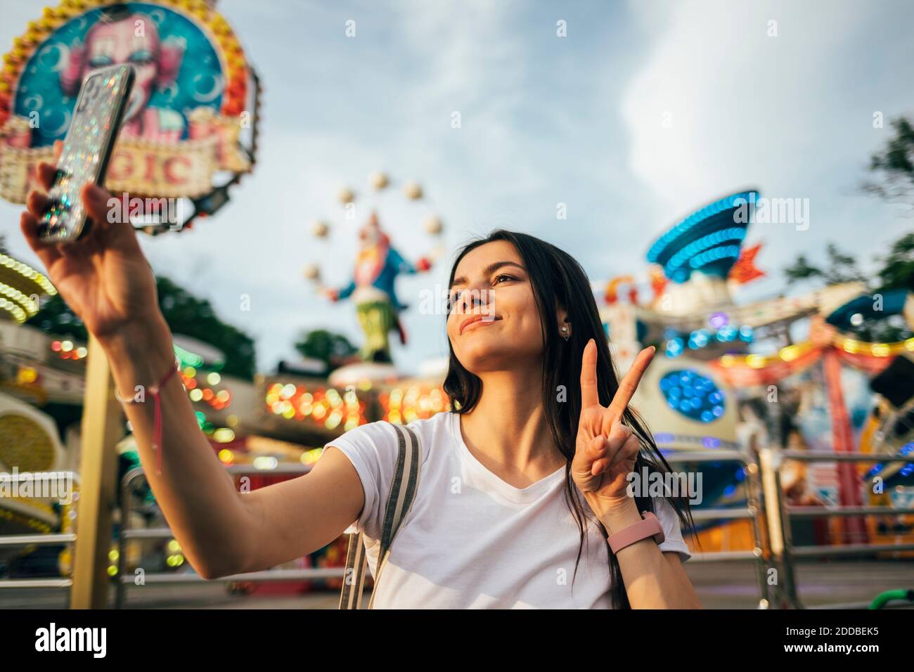 Beautiful young woman taking selfie through smart phone while gesturing peace sign at amusement park during summer Stock Photo