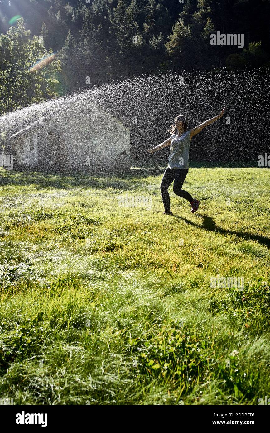 Woman standing with arms outstretched in front of sprinkler on grass during sunny day Stock Photo