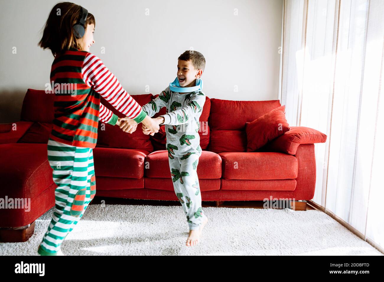 Brother and sister with headphones holding hands while dancing against sofa in living room Stock Photo
