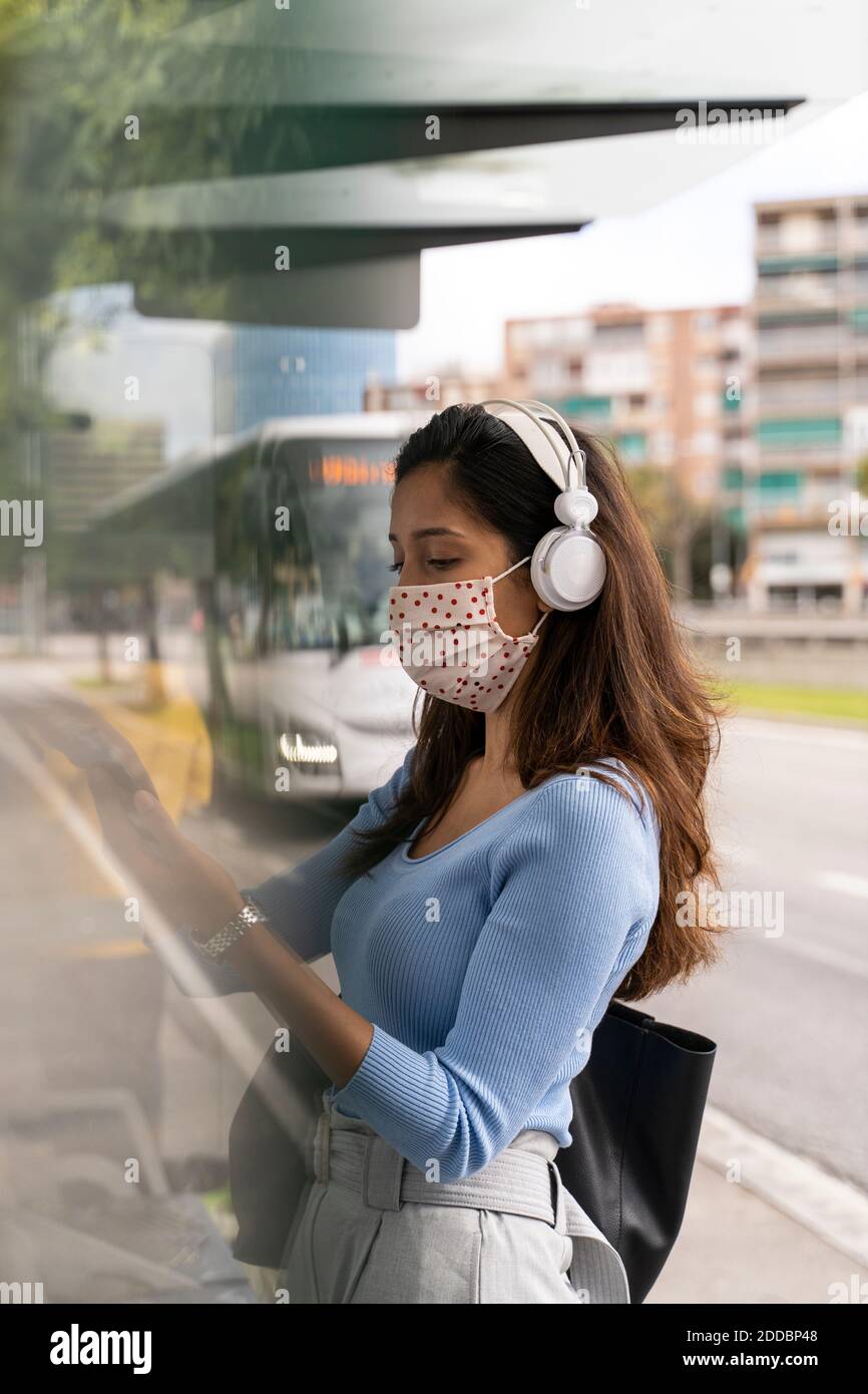 Woman in face mask wearing headphones while using smart phone at bus stand during COVID-19 Stock Photo
