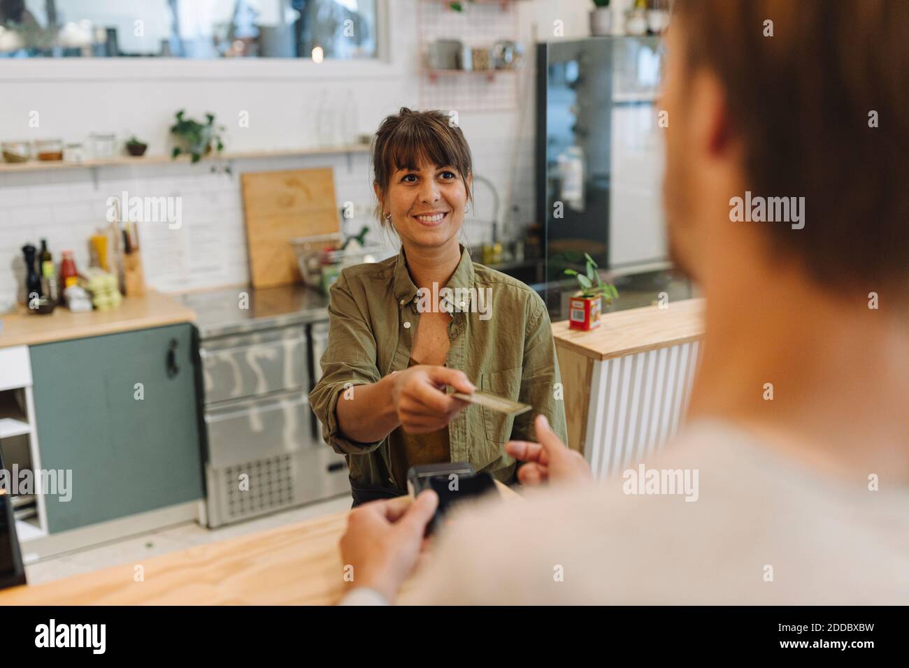 Smiling female owner giving back credit card to male customer at checkout in coffee shop Stock Photo
