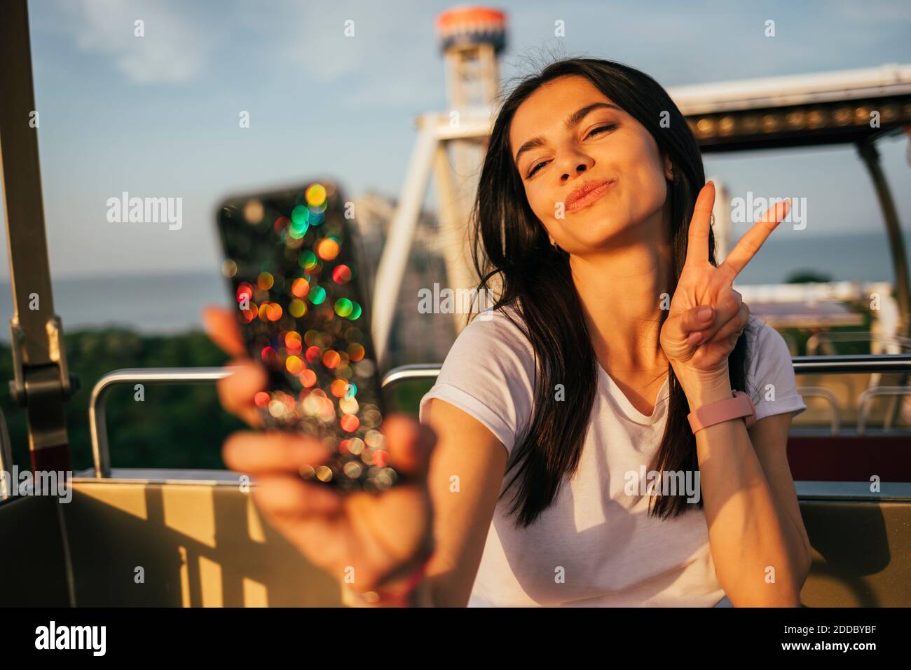 Beautiful young woman taking selfie through mobile phone while puckering and gesturing peace sign on Ferris wheel Stock Photo