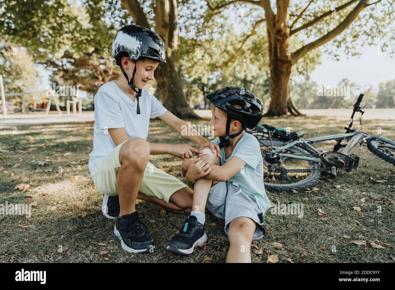 Boy smiling after putting bandage on younger brother knee sitting in public park Stock Photo
