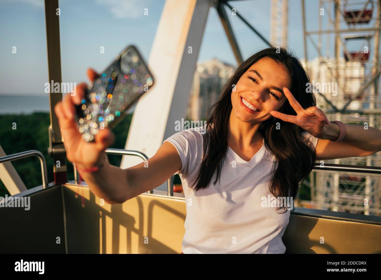 Cheerful young woman gesturing peace sign while taking selfie on Ferris wheel at amusement park Stock Photo