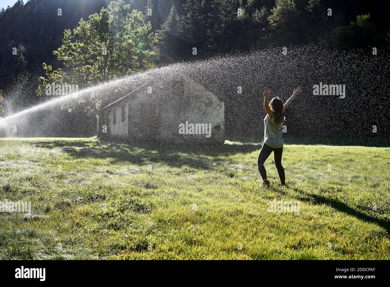 Woman standing in front of sprinkler on grass during sunny day Stock Photo