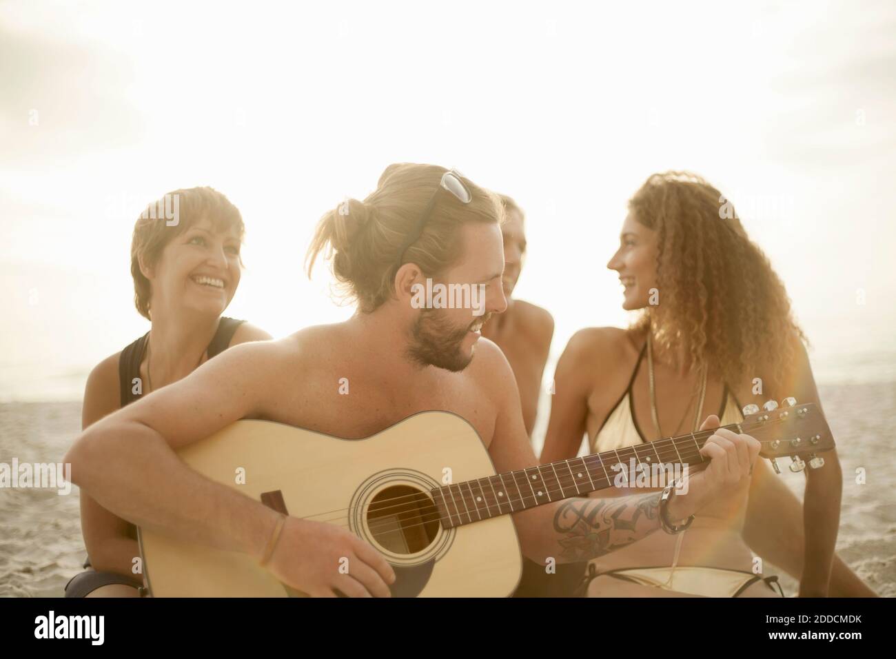 Mid adult man playing guitar while his friends singing sitting on sand at beach Stock Photo
