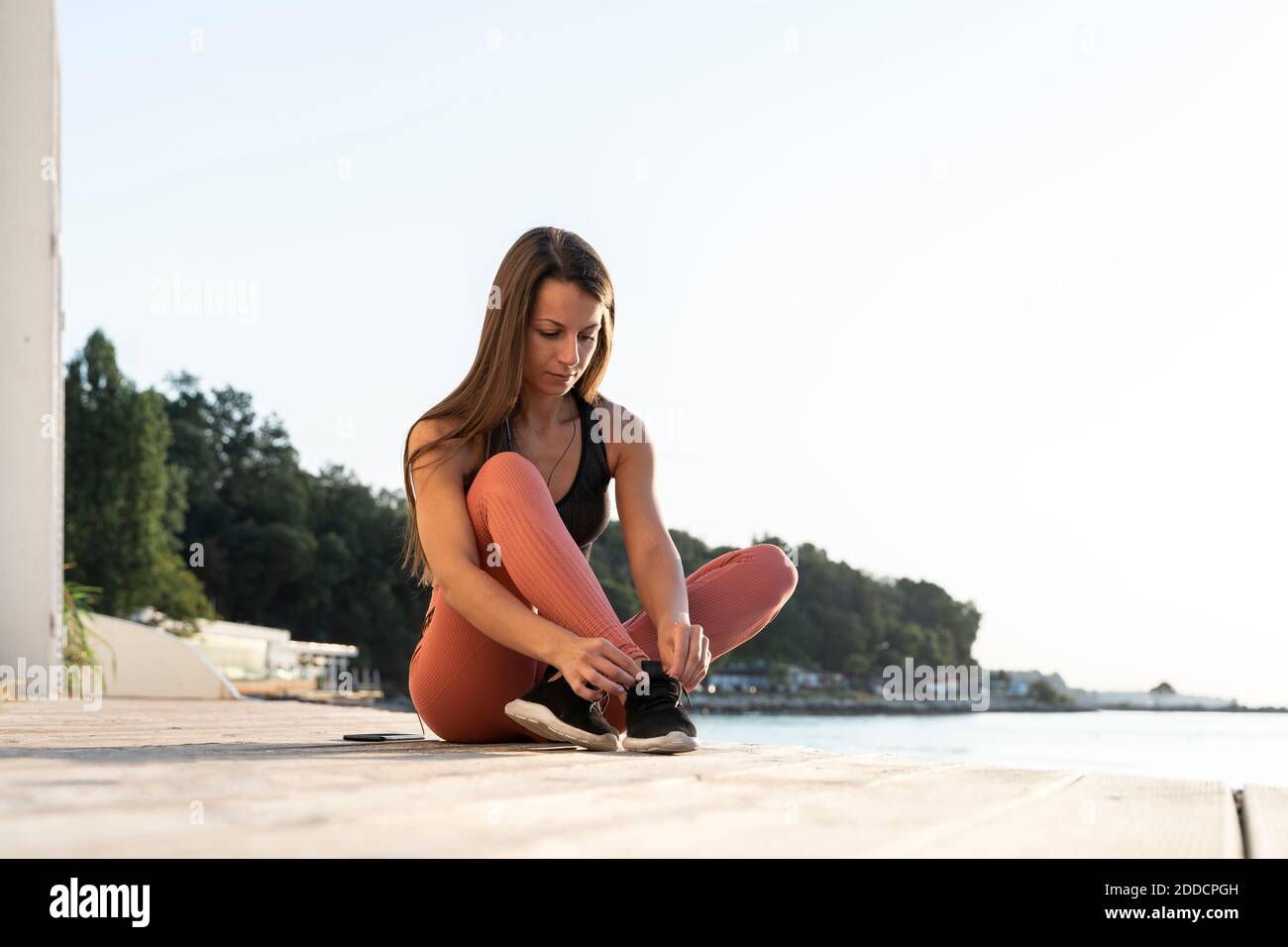 Sportswoman tying shoelace sitting on promenade by sea during sunrise Stock Photo