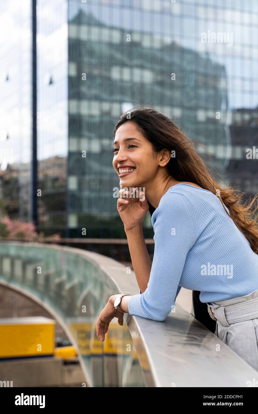 Thoughtful businesswoman smiling while leaning on railing of footbridge Stock Photo