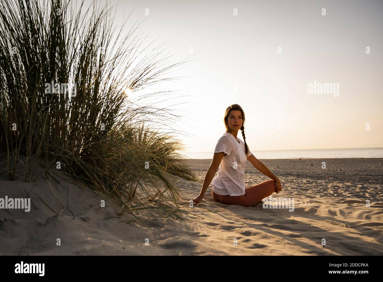 Beautiful young woman practicing yoga while sitting by plant at beach against clear sky during sunset Stock Photo