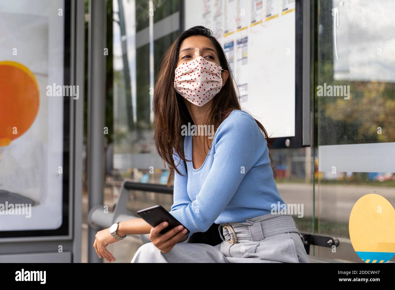 Woman with smart phone wearing face mask while waiting at bus stand during COVID-19 Stock Photo