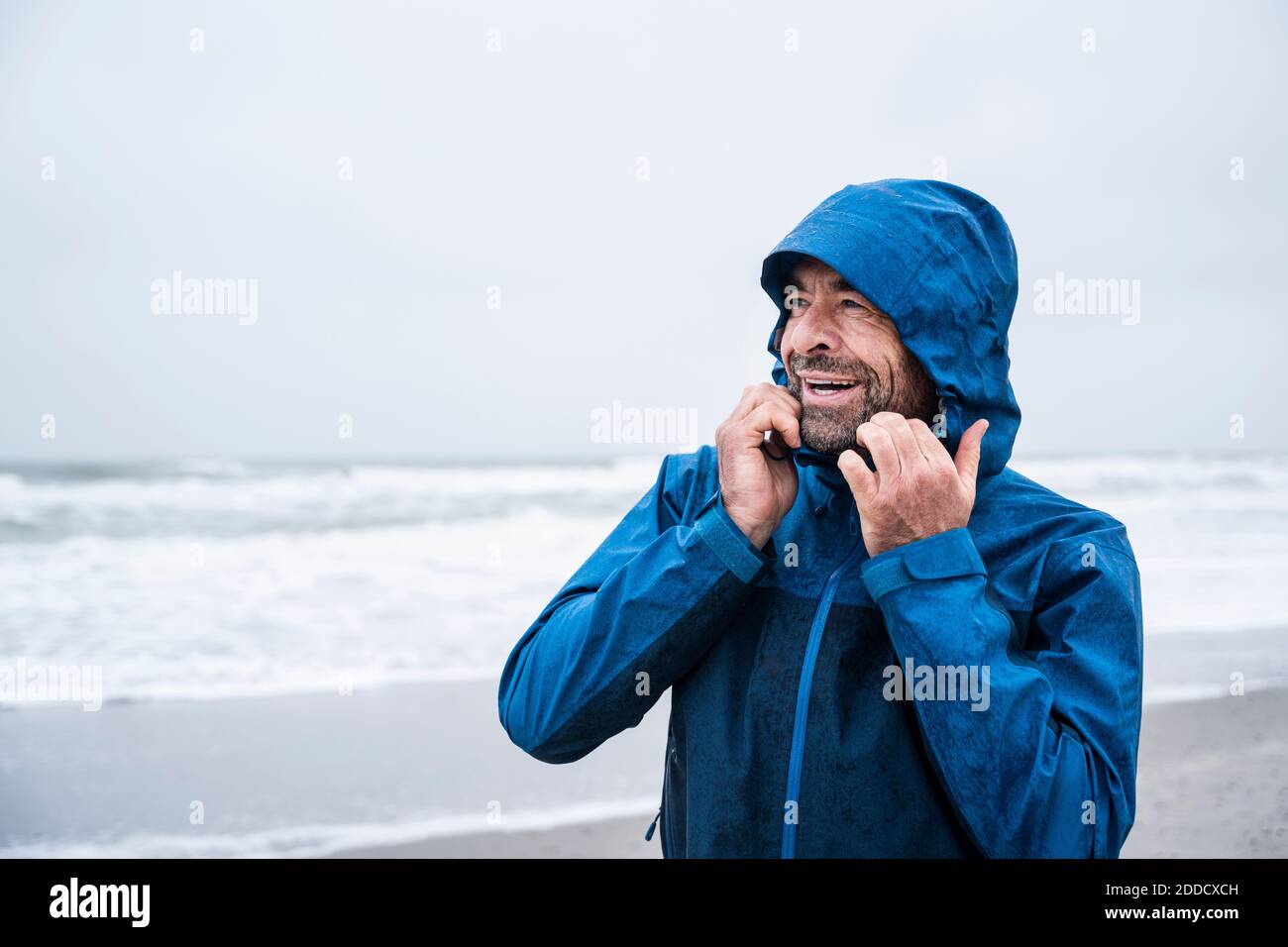 Happy mature man in blue raincoat looking away while standing at beach Stock Photo