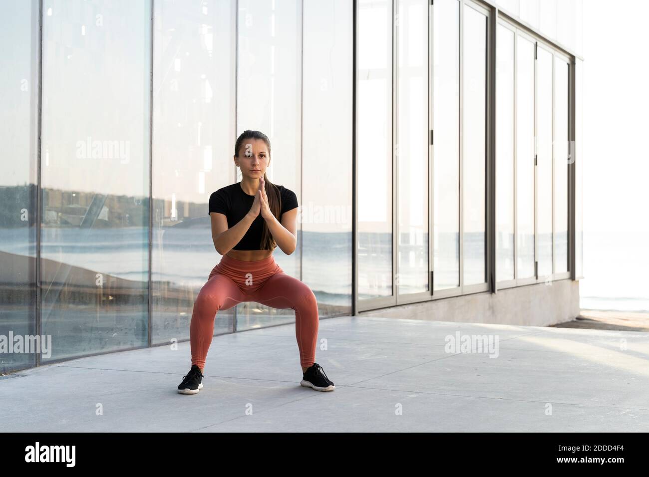 Young sportswoman doing squat exercise on promenade against glass window Stock Photo