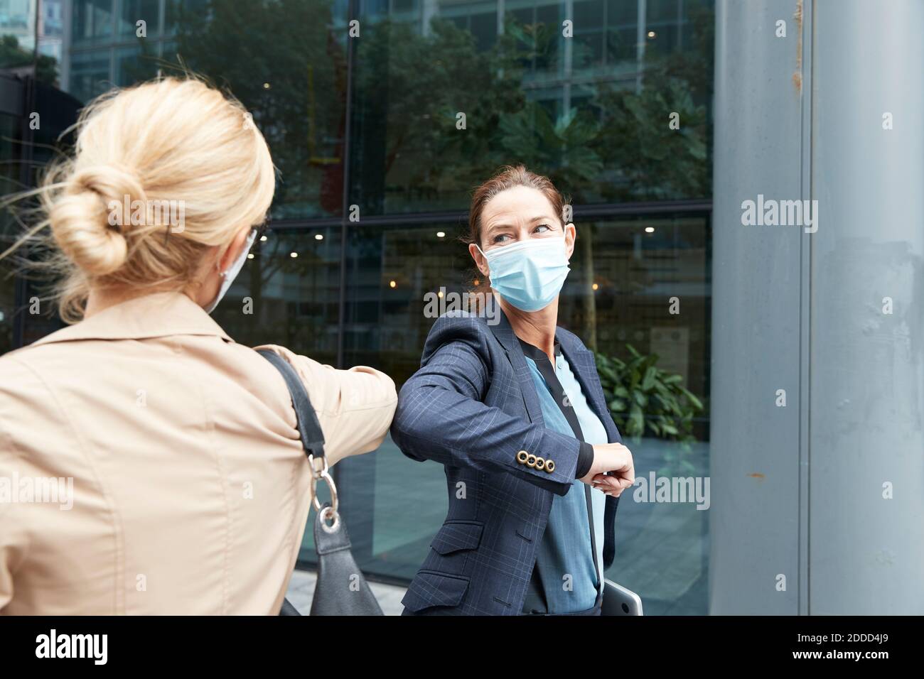 Businesswoman and colleague with face mask greeting with elbow bump while standing against office building Stock Photo