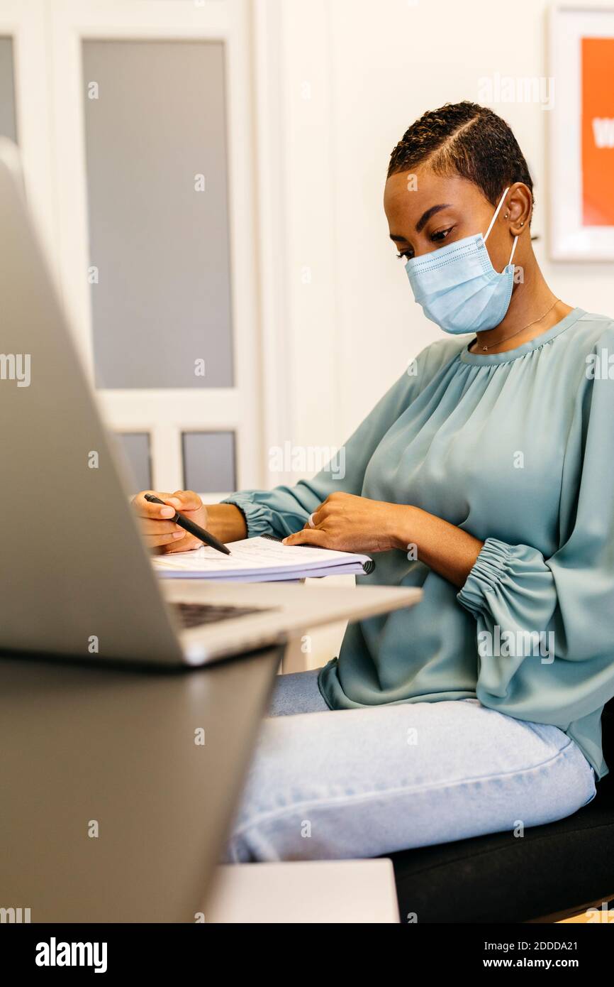 African businesswoman with safety mask reading papers while sitting in ...